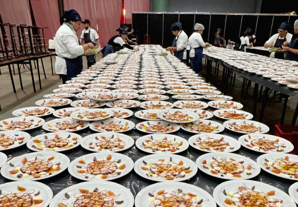 La imagen ilustra la calidad gastronómica del catering mostrando hileras de platos con un cuidado carpaccio de pulpo.